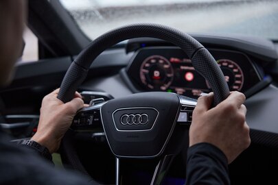 Person holding an Audi steering wheel with digital display in background.