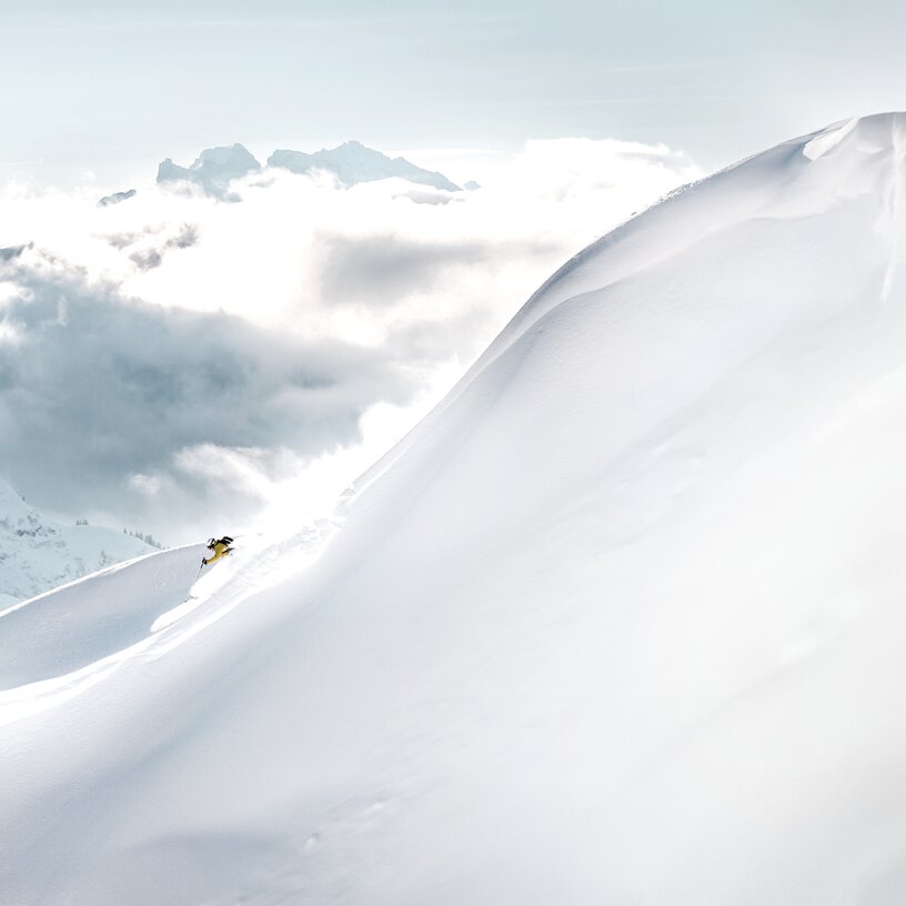 A skier on a snowy, high mountain with clouds in the background.