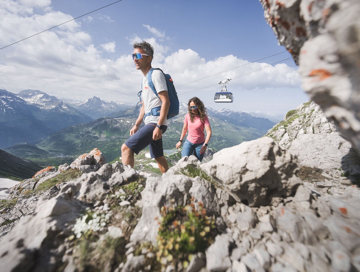 Two people hiking on a rocky mountain trail with a cable car in the background.