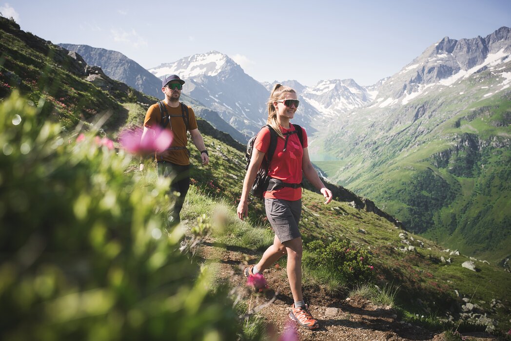 Zwei Personen wandern durch die grüne Natur in den Bergen von St. Anton am Arlberg mit schneebedeckten Gipfeln im Hintergrund.