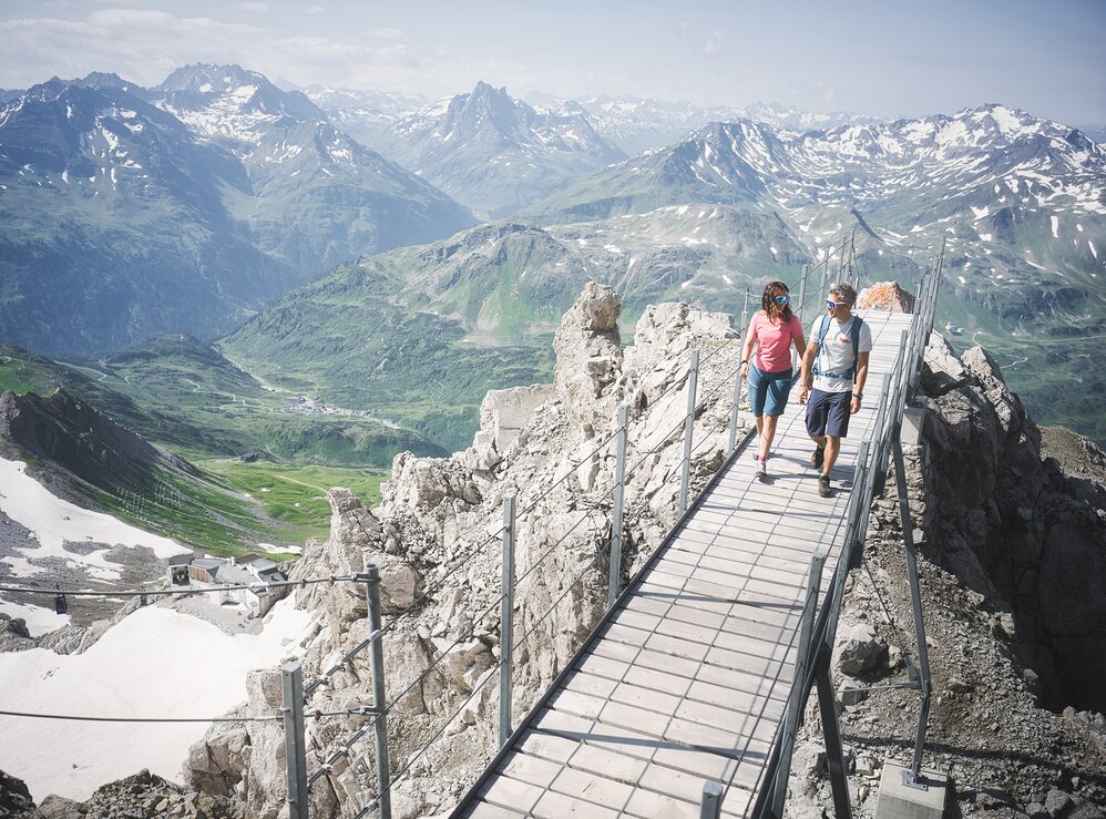 Two people walking on a narrow bridge in the mountains with snow-capped peaks in the background.