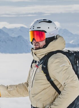 A skier with a helmet and backpack in front of snowy mountains.