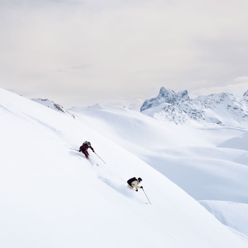 Two skiers dynamically gliding through untouched powder snow with a stunning mountain backdrop.