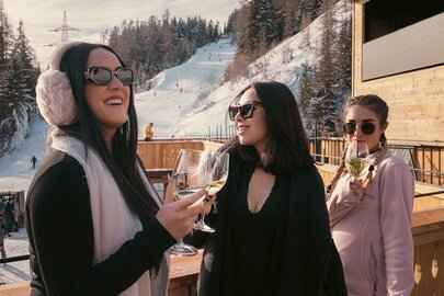 Drei schön gekleidete Frauen genießen Getränke auf einer Terrasse im Schnee.