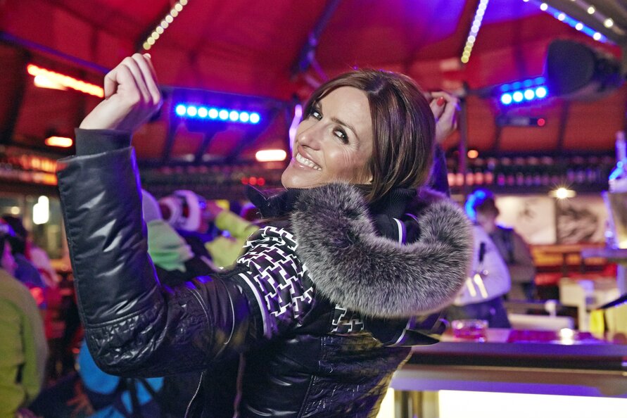 Frau in Winterjacke lächelt und hebt beim Tanzen die Arme auf einer Party.