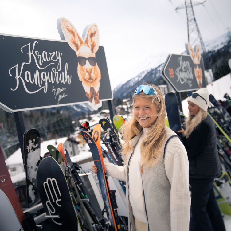 Woman in ski gear smiling in front of skis at Krazy Kanguruh, snowy mountains in background.
