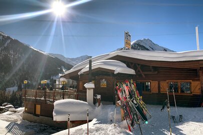 Wooden hut under bright sunshine in snowy mountains, with skis and poles in the foreground.