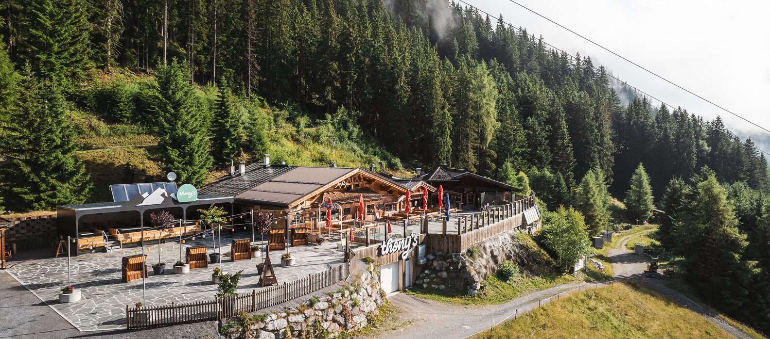 A mountain lodge nestled in a forest with terrace and umbrellas.