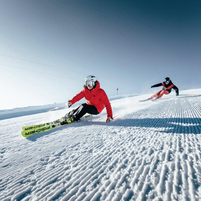 Zwei Skifahrer fahren auf einer frisch präparierter Piste den Berg hinunter.