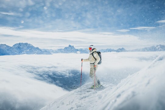 Ein Freerider steht auf einem schneebedeckten Berg mit Skistöcken in der Hand, umgeben von Wolken und Bergen.