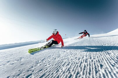 Two skiers gracefully glide over a freshly groomed slope in sunshine.