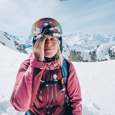 Eine Frau in Skiausrüstung wischt sich mit der rechten Hand den Schnee aus dem Gesicht.