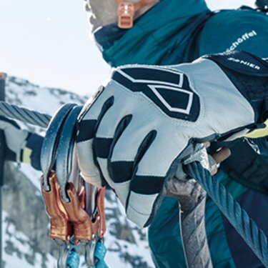Ein Kletterer mit Helm, Handschuhen und Skiern am Rücken erklimmt am Seil einen verschneiten Bergpfad.