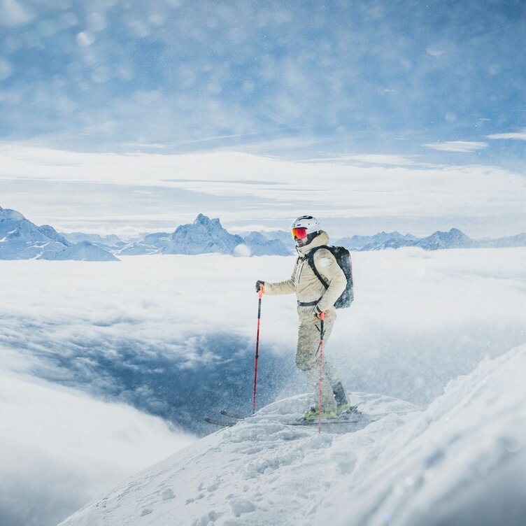 Ein Skifahrer steht auf einem schneebedeckten Berg mit Blick auf ein Wolkenmeer.