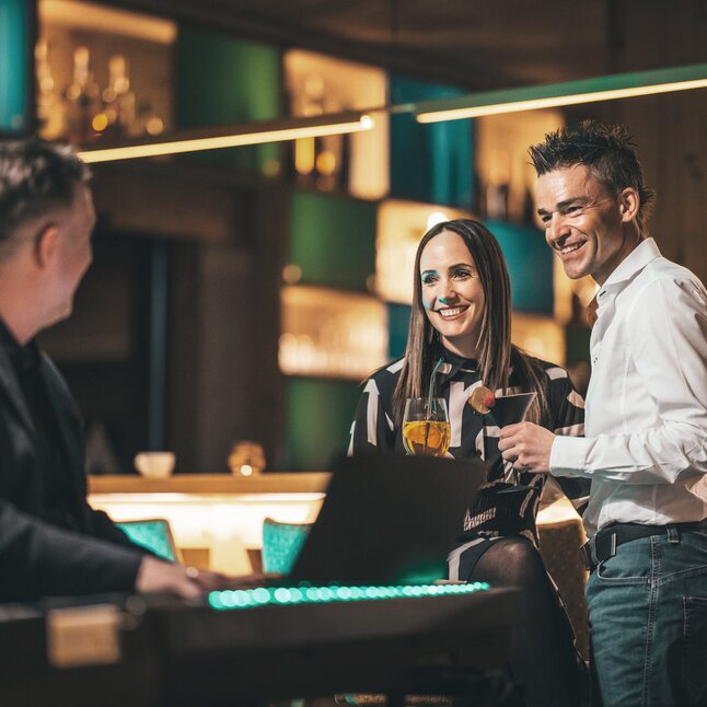 Ein Paar steht mit Getränken in der Hand an der Bar und unterhält sich mit einer dritten Person.