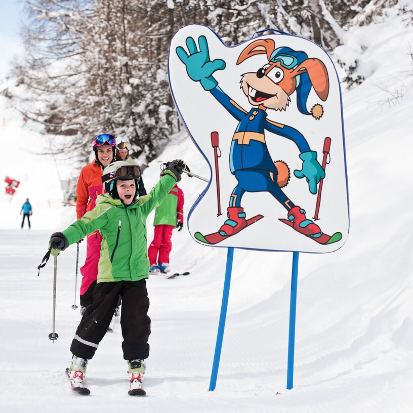 Kind und Erwachsener auf Skiern neben einem Hasen-Maskottchen.