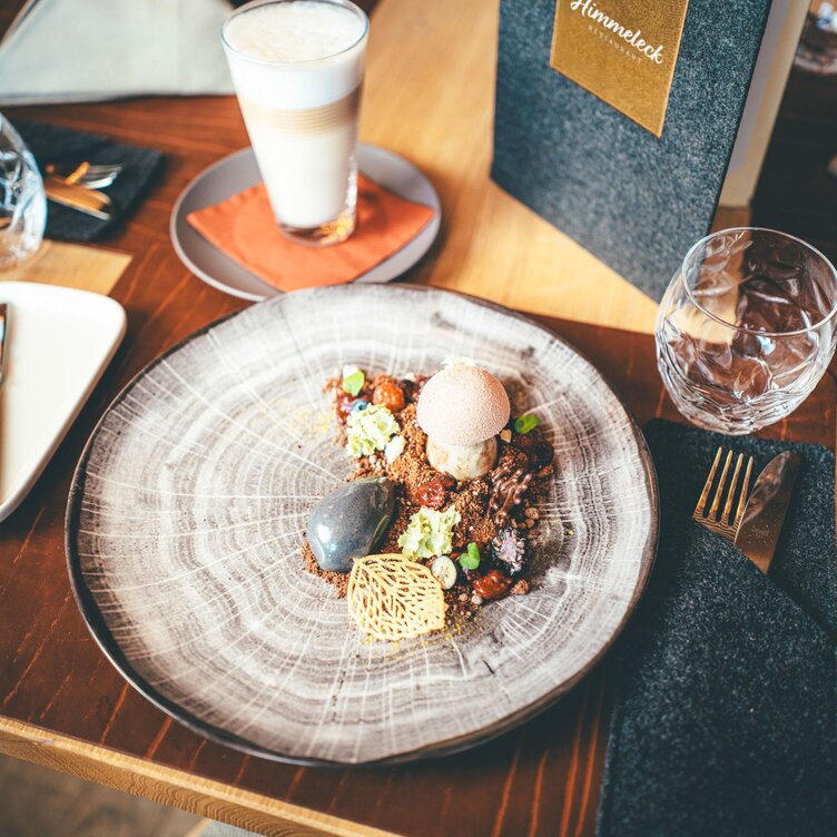 A creatively presented dessert on a plate, next to a menu labeled 'Himmeleck'. In the background, a latte macchiato.