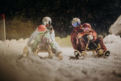 Eine Familie bestehend aus zwei Erwachsenen und zwei Kindern rodeln abends auf einer verschneiten Piste.