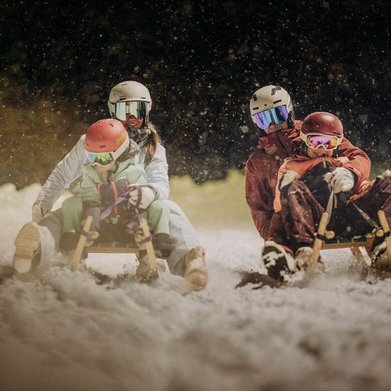 Four people tobogganing on a snowy slope at night.