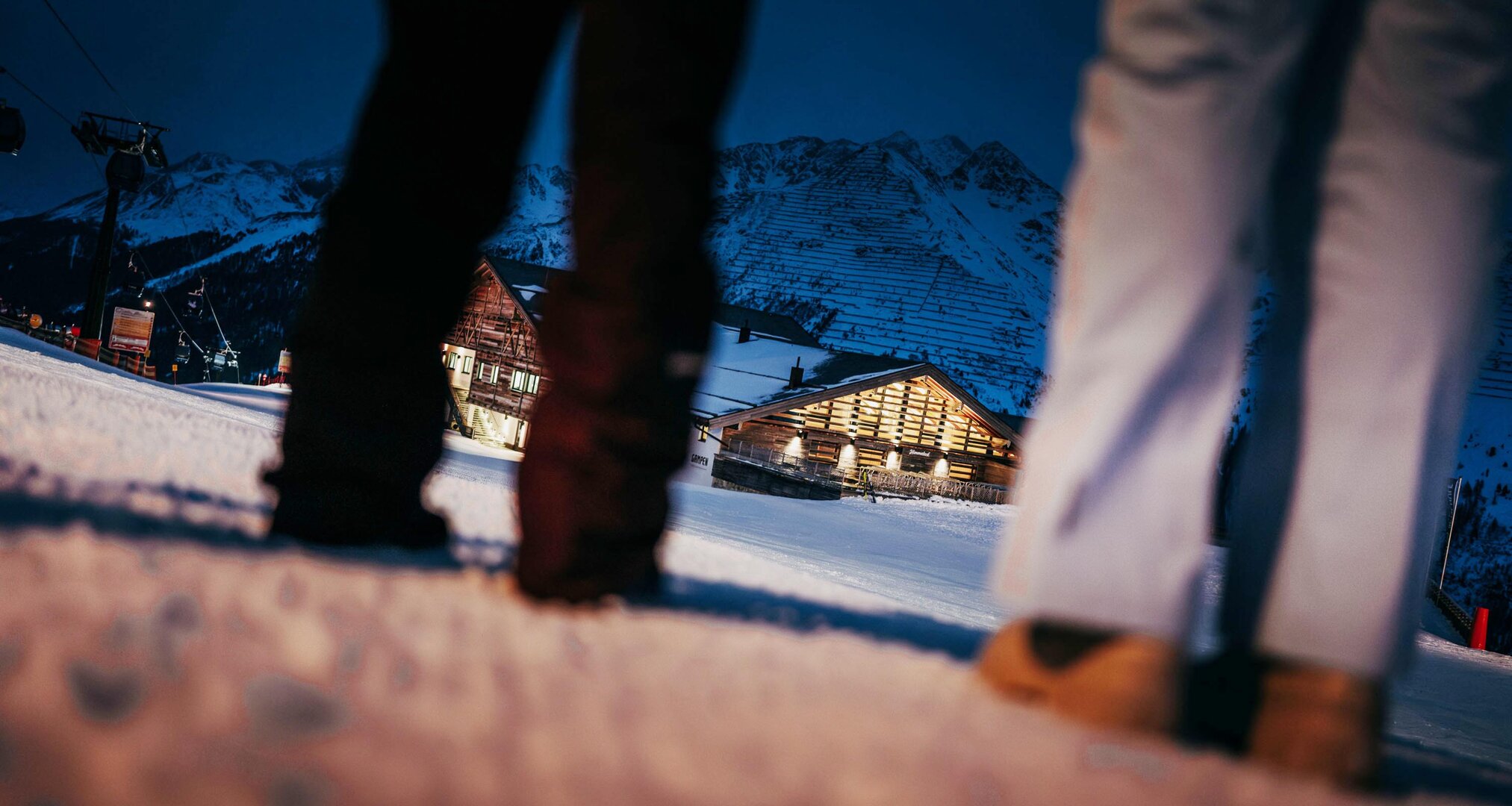 Zwei Personen in Winterkleidung stehen vor einer beleuchteten Berghütte im Schnee.