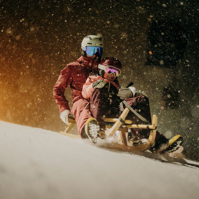 Zwei Personen rodeln nachts im Schneefall auf einem Holzschlitten.