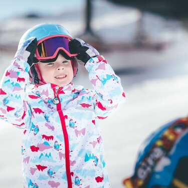 Child wearing colorful ski jacket and goggles on snowy mountain.