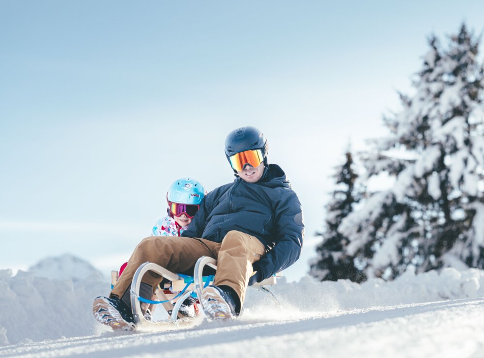 Familie beim Rodeln im Schnee mit Schlitten, Helm und Brille.