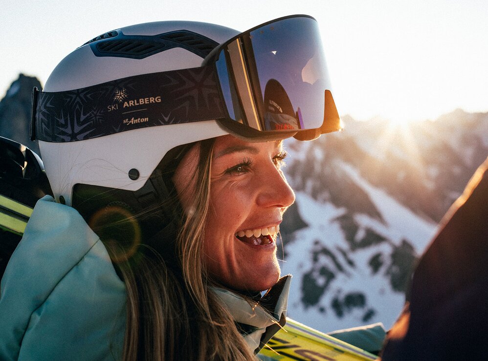 Lächelnde Frau mit Skihelm bei Sonnenuntergang in den Bergen.