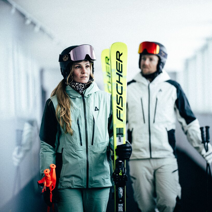 Two people with skis and gear in a hallway.