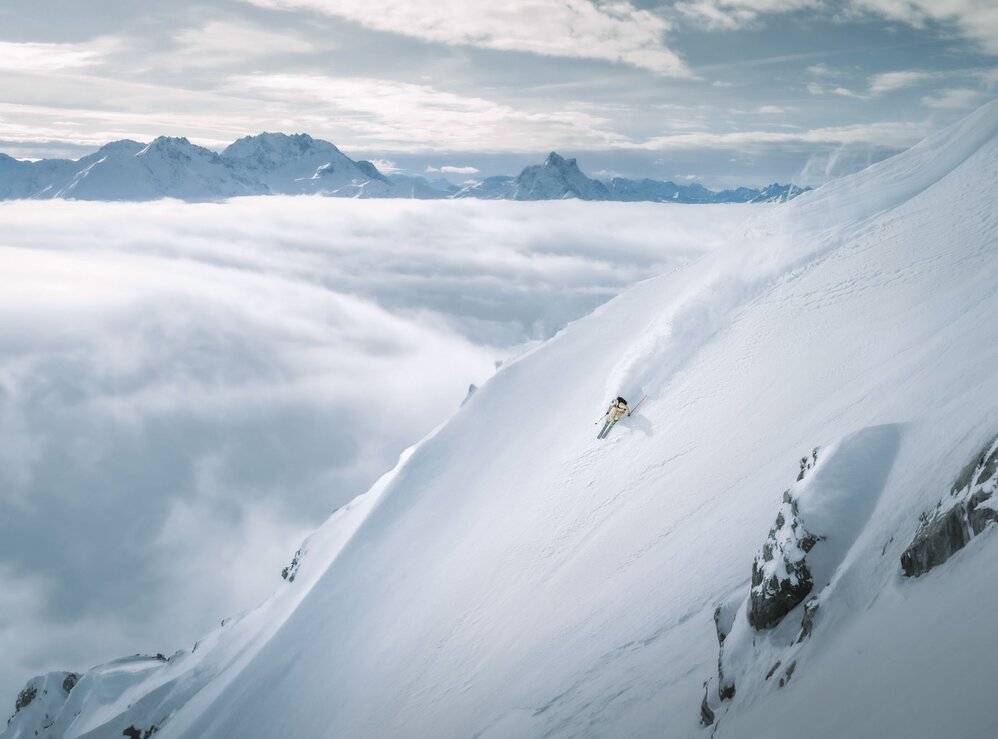 A skier descends a steep slope in the St. Anton am Arlberg ski area, surrounded by clouds and mountain peaks.
