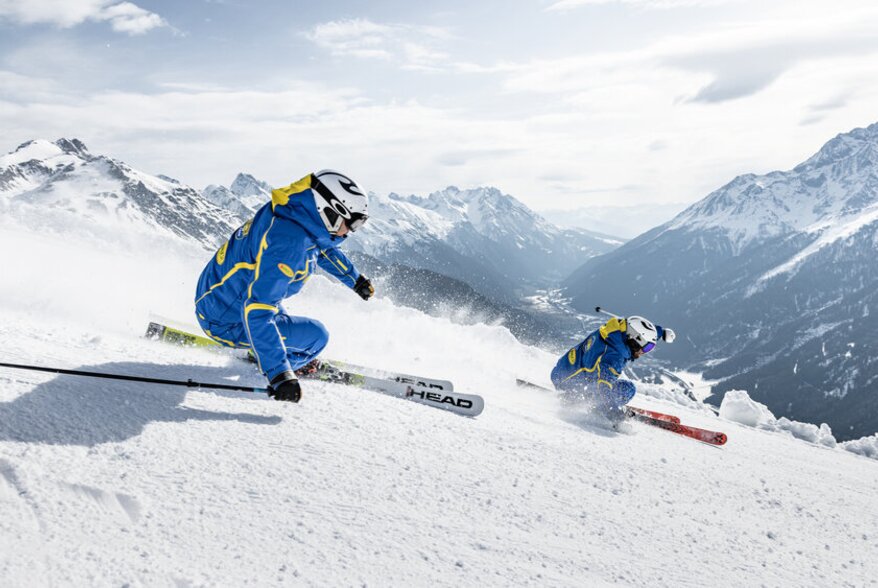 Zwei Skifahrer in blauen Jacken fahren schnell die verschneite Piste hinunter.