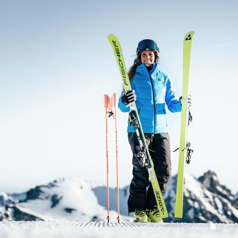 A woman stands on a slope with skis and a helmet, mountains in the background.
