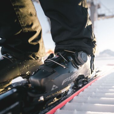 Close-up of a ski boot on snow with a cable car gondola in the background.