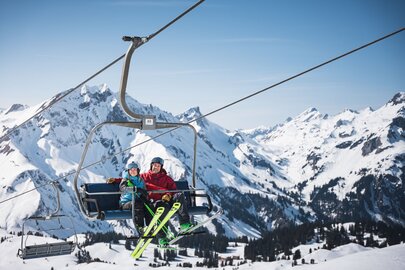 Zwei Personen auf einem Sessellift am Arlberg, umgeben von schneebedeckten Bergen.