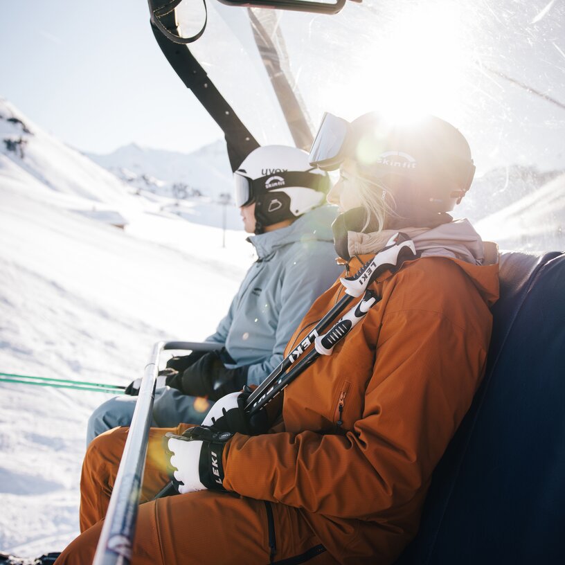Zwei Skifahrer mit orangefarbener und grauer Kleidung auf einem Sessellift im Schnee.