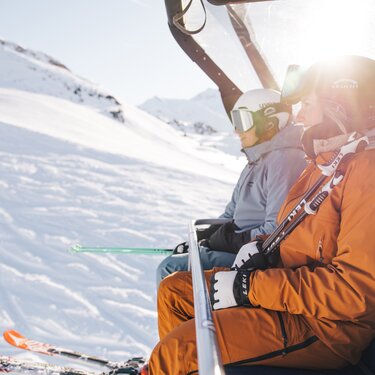 Zwei Skifahrer auf einem Sessellift in schneebedeckten Bergen.