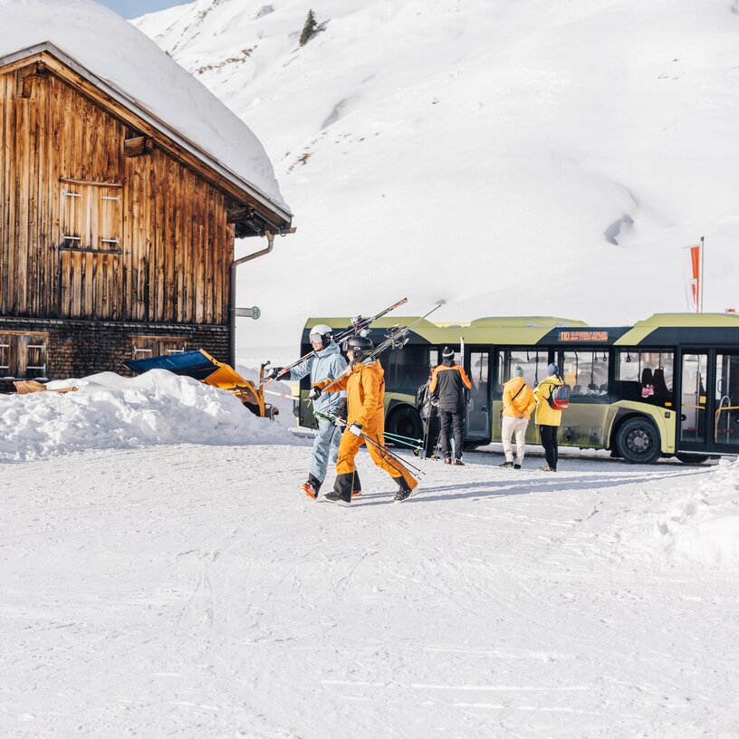 Skiers in front of a shuttle bus in a snowy mountain landscape.