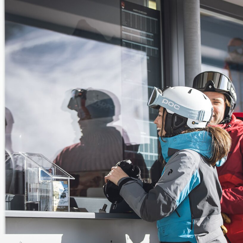 Zwei Skifahrer im Gespräch mit einem Ticketverkäufer am Schalter.
