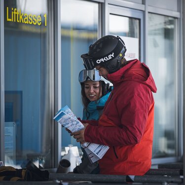 Two people in ski gear, holding a map at a ticket booth.