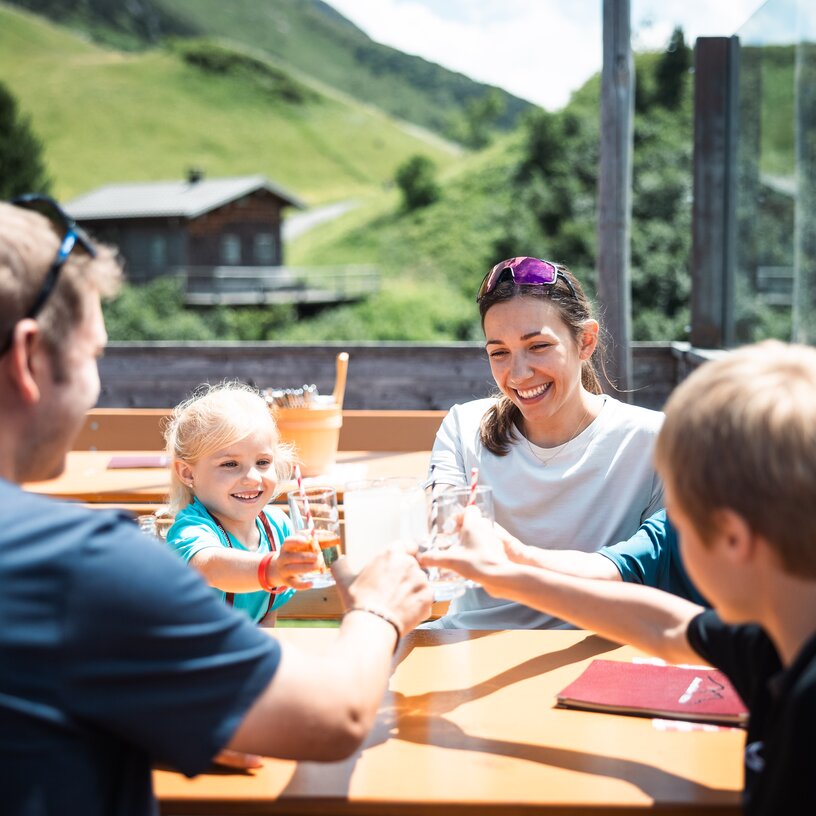 Eine lächelnde Familie stößt draußen an einem Tisch an, umgeben von Bergen.