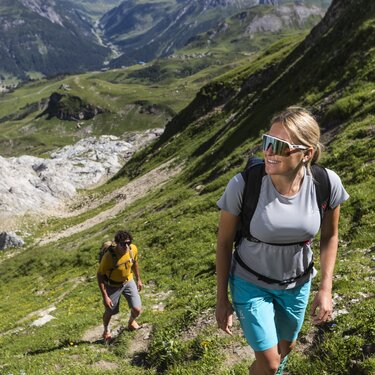 Zwei Personen wandern einen grünen Bergpfad entlang.