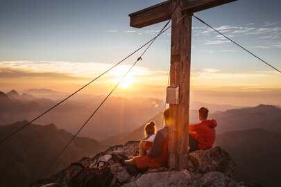 Two people sitting on a mountain at sunrise, next to a summit cross.