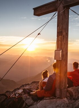 Two people sitting on a mountain at sunrise, next to a summit cross.