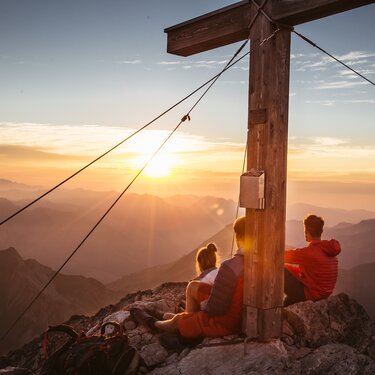 Zwei Personen sitzen bei Sonnenaufgang auf einem Berg neben einem Gipfelkreuz.