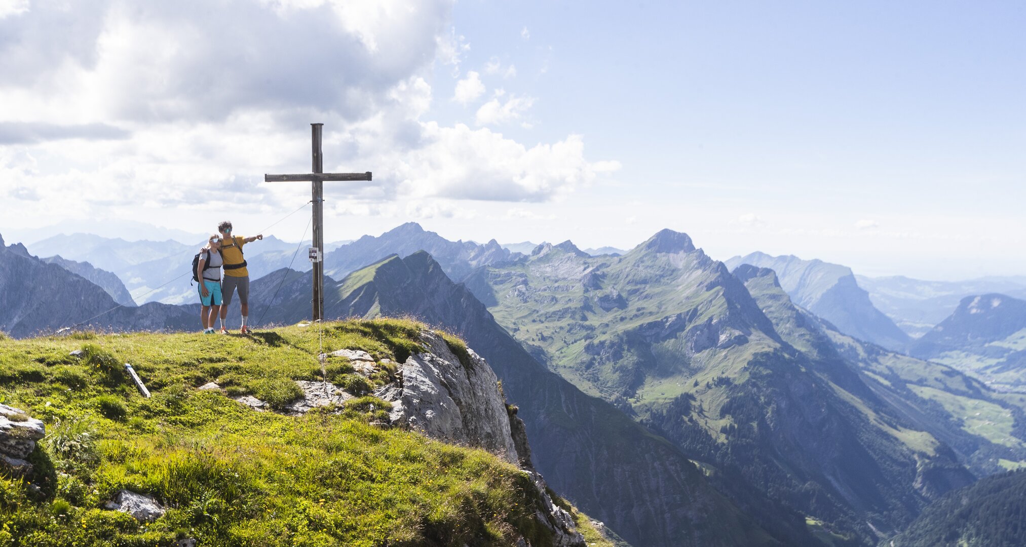 Two people with backpacks on mountain peak beside wooden cross, surrounded by scenic mountain panorama.