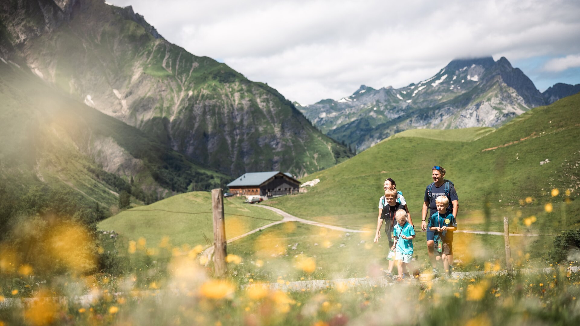 Familie wandert entlang eines Pfades über blühende Almwiesen, im Hintergrund ist eine Hütte zu sehen.