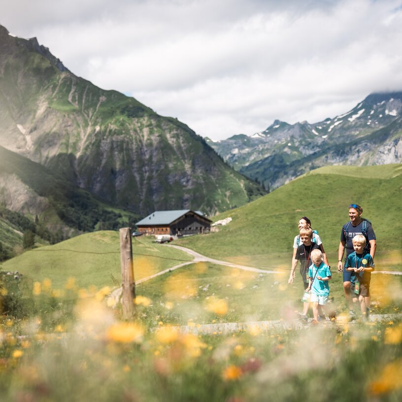 Familie wandert entlang eines Pfades über blühende Almwiesen, im Hintergrund ist eine Hütte zu sehen.