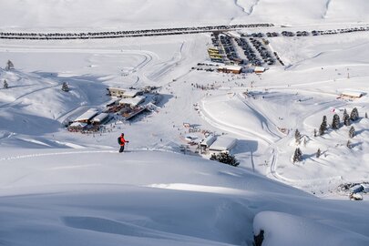 Skigebiet mit Schnee, Skifahrer und Parkplatz im Hintergrund.