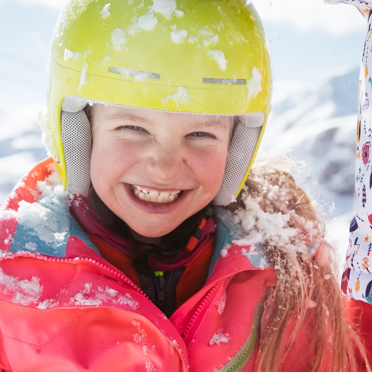 Ein lachendes Kind mit gelbem Skihelm und einer pinken Skijacke im Schnee.