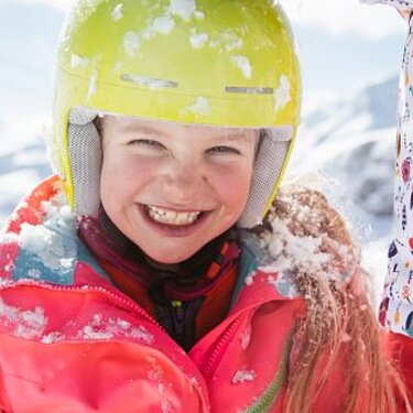 Ein Mädchen mit gelbem Skihelm und pinker Jacke lächelt im Schnee.